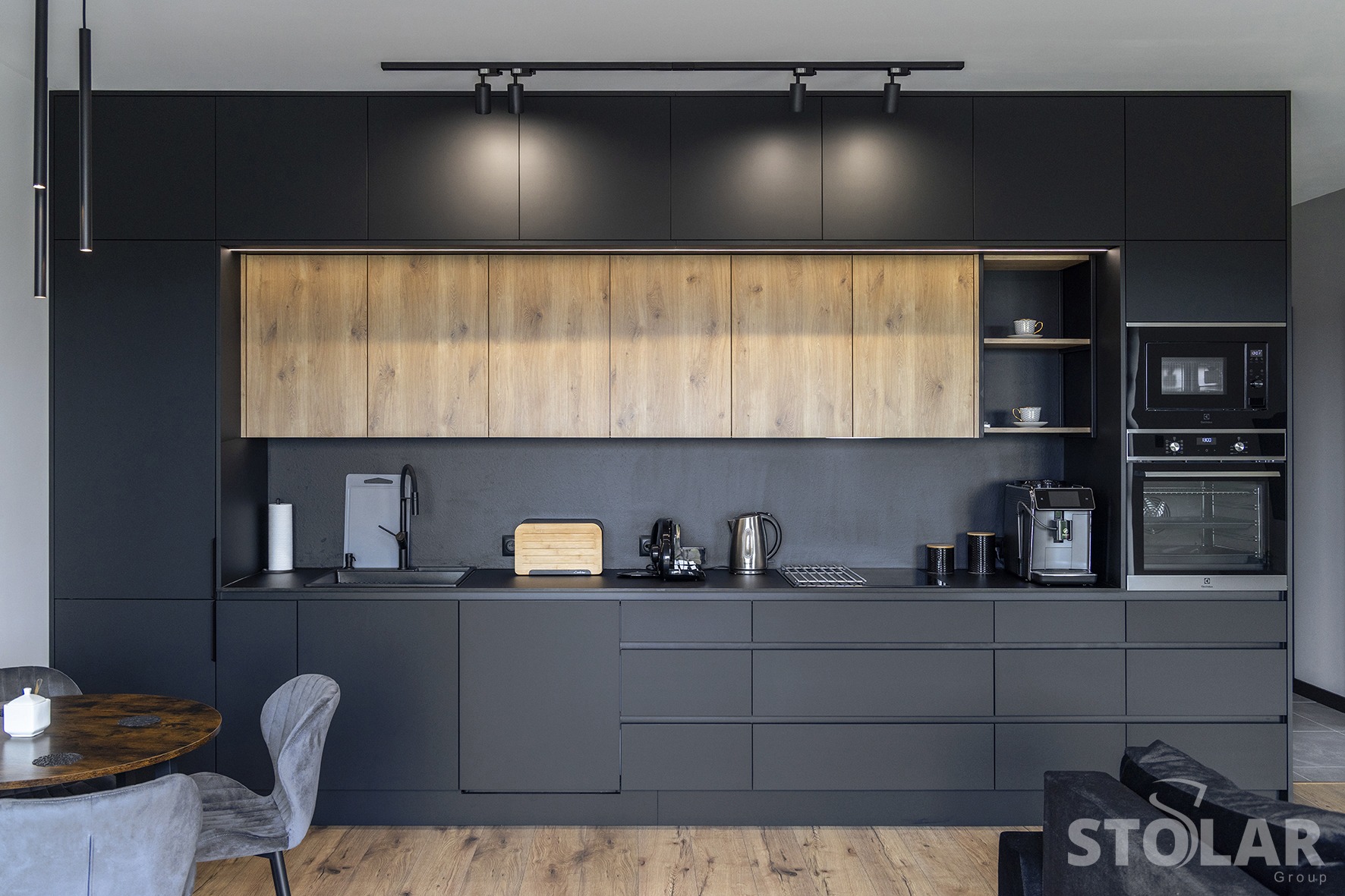 Nowoczesna kuchnia w odcieniach szarości i drewna z oświetleniem punktowym, ekspres do kawy i piekarnik w zabudowie, widok na stół z krzesłami i fragment sofy.