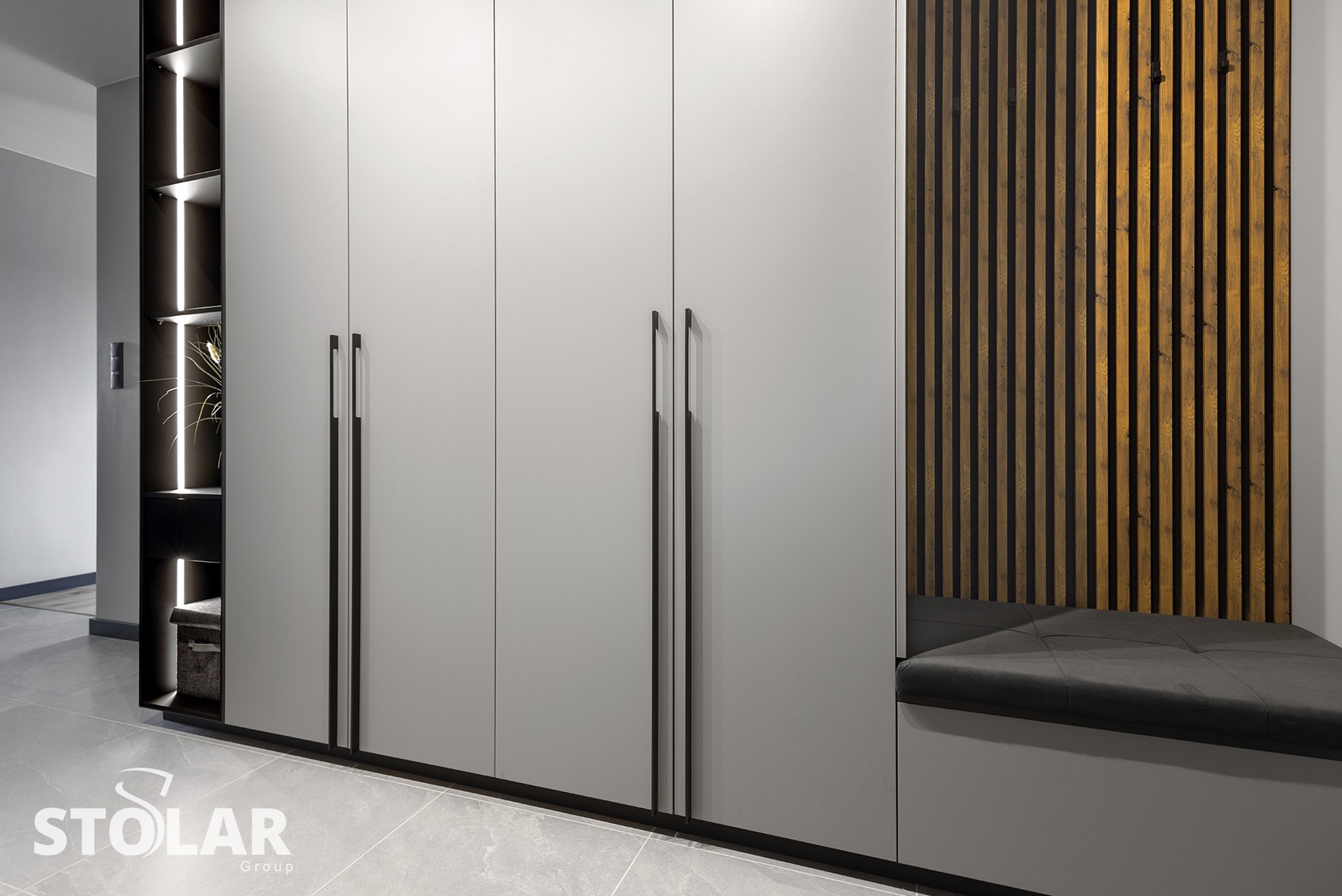 Nowoczesna zabudowa przedpokoju z szarymi szafami z długimi, czarnymi uchwytami, otwartym regałem z podświetleniem LED i siedziskiem z drewnianą ścianką z lameli.