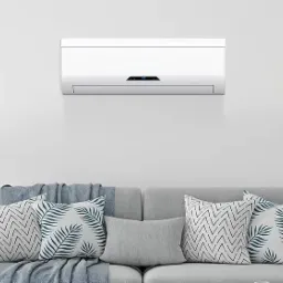 Biały klimatyzator ścienny z wyświetlaczem temperatury, zamontowany nad szarą sofą z dekoracyjnymi poduszkami w salonie z lampą w tle.