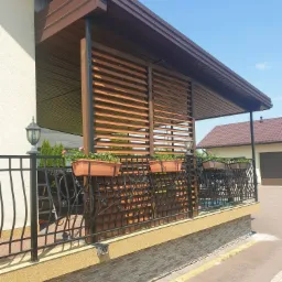 Zadaszony taras z drewnianymi lamelami, zdobiony kutą balustradą z donicami pełnymi kwiatów, oświetlony ozdobnymi lampami, widok z zewnątrz.