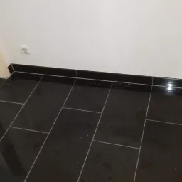 Błyszcząca, czarna podłoga wyłożona prostokątnymi płytkami, widoczny biały cokół i fragment białej ściany z gniazdkiem elektrycznym, oraz kartonowe pudło w tle.