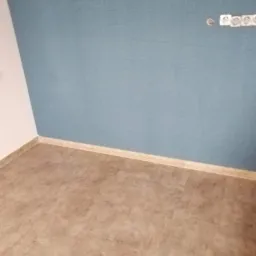 Pusty pokój po remoncie: błękitna ściana z gładzią, brązowe panele podłogowe, listwa przypodłogowa w kolorze drewna, gniazdka elektryczne na ścianie.