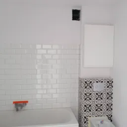 Wykończona łazienka z białą wanną obłożoną białymi, błyszczącymi płytkami typu metro, obudowa stelaża WC i fragment podłogi wykończone czarno-białymi płytkami w marokański wzór, wisząca szafka...