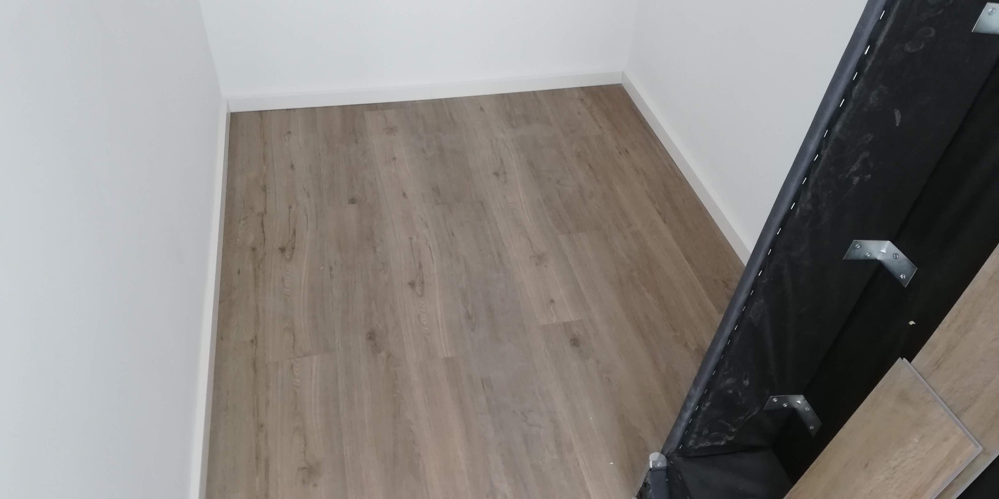 Nowo ułożone panele podłogowe w jasnym odcieniu drewna, widoczne fragmenty ściany pomalowanej na biało i element konstrukcyjny z metalowymi mocowaniami w prawym dolnym rogu.