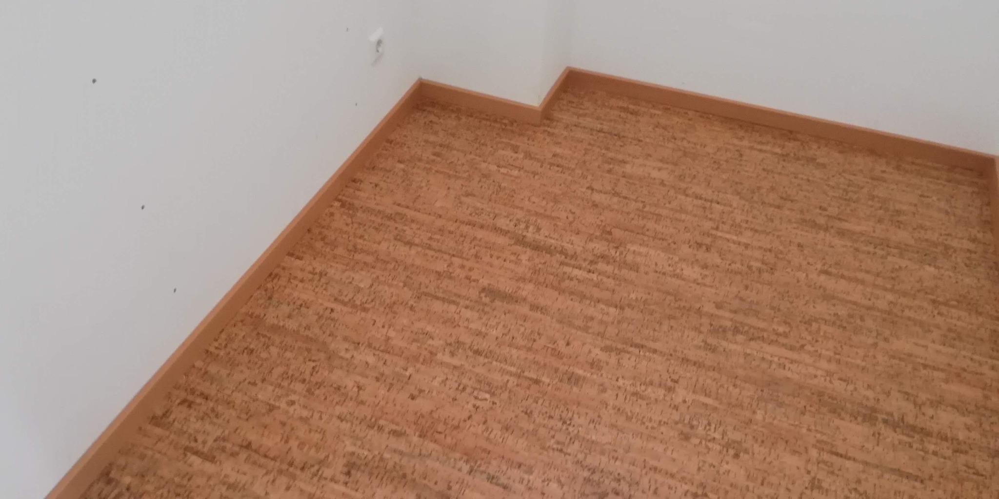 Nowo ułożona podłoga korkowa z jasnymi listwami przypodłogowymi w narożniku pokoju, widoczne ślady po wierceniu na ścianie.