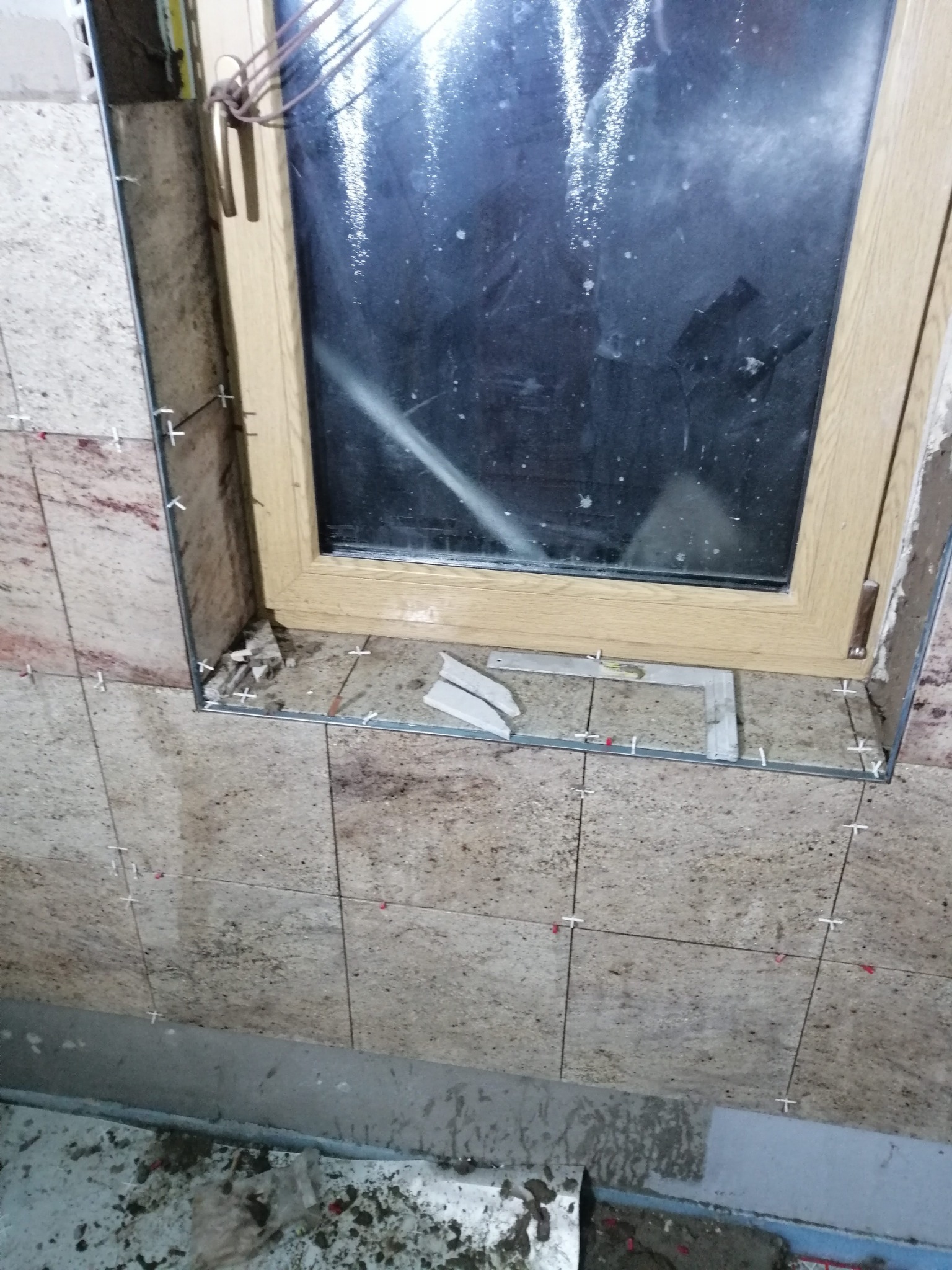 Montaż płytek granitowych wokół okna w łazience, widoczne krzyżyki dystansowe i aluminiowe profile wykończeniowe.