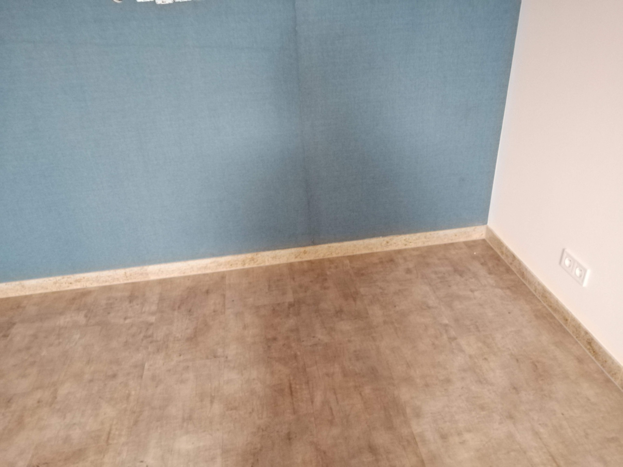 Wnętrze po remoncie: Ściana pomalowana na niebiesko, panele podłogowe w odcieniach brązu, widoczne listwy przypodłogowe i gniazdka elektryczne.