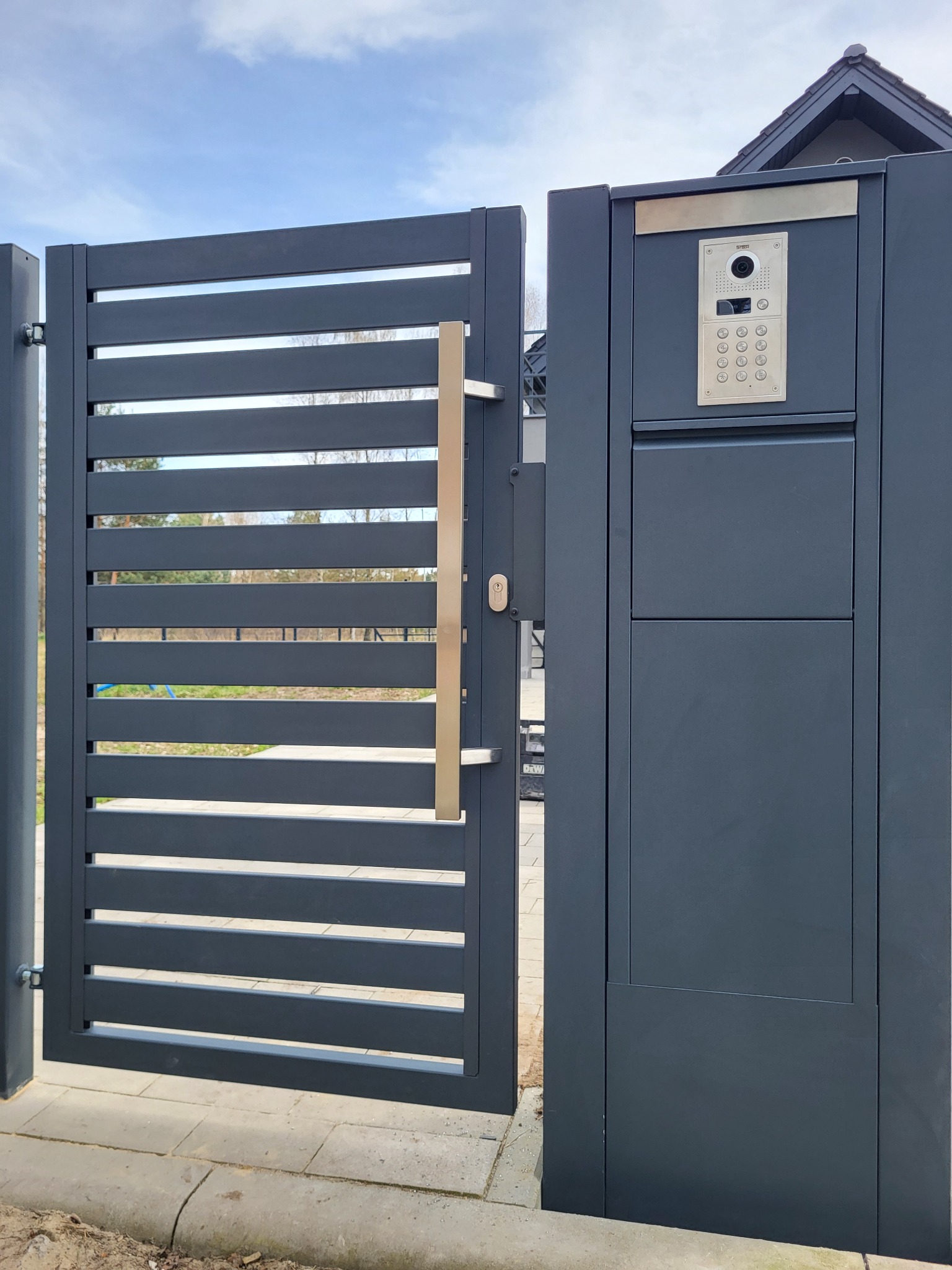 Nowoczesna, antracytowa brama wjazdowa z poziomymi panelami, długim, pionowym uchwytem ze stali nierdzewnej i zintegrowanym wideodomofonem.