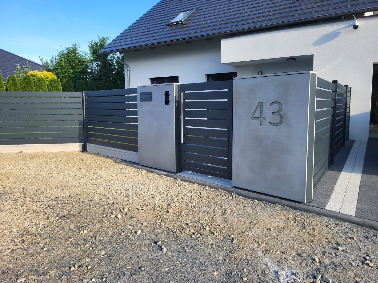 Nowoczesna brama wjazdowa i przęsła ogrodzeniowe z poziomych profili w kolorze grafitowym, zamontowane na betonowych słupkach z numerem domu 43, widok z zewnątrz posesji.