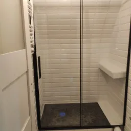 Nowoczesna kabina prysznicowa z czarną ramą i przesuwnymi drzwiami, wyłożona białymi, cegiełkowymi płytkami, z siedziskiem wewnątrz i ciemną podłogą z niebieskim odpływem, na podłodze wzorzyste...