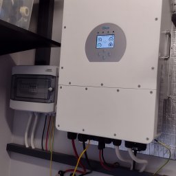 NordInstal Wojciech Dobrzański - Biały inwerter solarny marki Deye zamontowany na ścianie, z widocznym panelem sterowania i podłączonymi kablami. Obok skrzynka bezpiecznikowa Noark i akumulator litowo-jonowy Deye. Widoczne...
