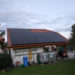 NordInstal Wojciech Dobrzański - Panele fotowoltaiczne na dachu budynku gospodarczego z czerwoną dachówką, widok na ogród z narzędziami i zbiornikami na wodę.