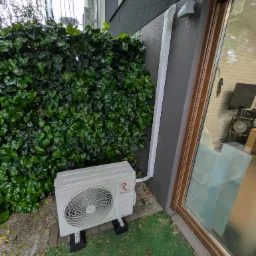 Biała jednostka zewnętrzna klimatyzacji Rotenso na zielonej trawie, obok ściany budynku z rurą odprowadzającą. Tło stanowi ściana z zielonego bluszczu.