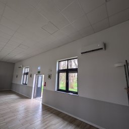 Dobre Instalacje - Wnętrze sali z klimatyzatorem na ścianie, widok na okna i drzwi. Podłoga wyłożona płytkami imitującymi drewno. Widoczne elementy instalacji.