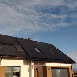 Dom jednorodzinny z ciemną dachówką i panelami fotowoltaicznymi, widoczne okna dachowe, maszt z polską flagą i numer domu 5, na tle błękitne niebo z chmurami.