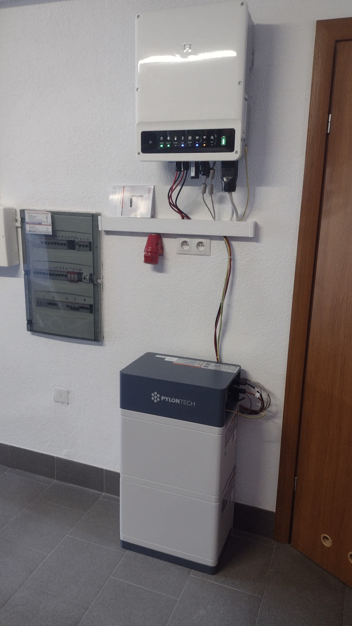 Biały inwerter fotowoltaiczny zawieszony na ścianie nad modułową baterią Pylontech, obok skrzynka z bezpiecznikami i brązowe drzwi.