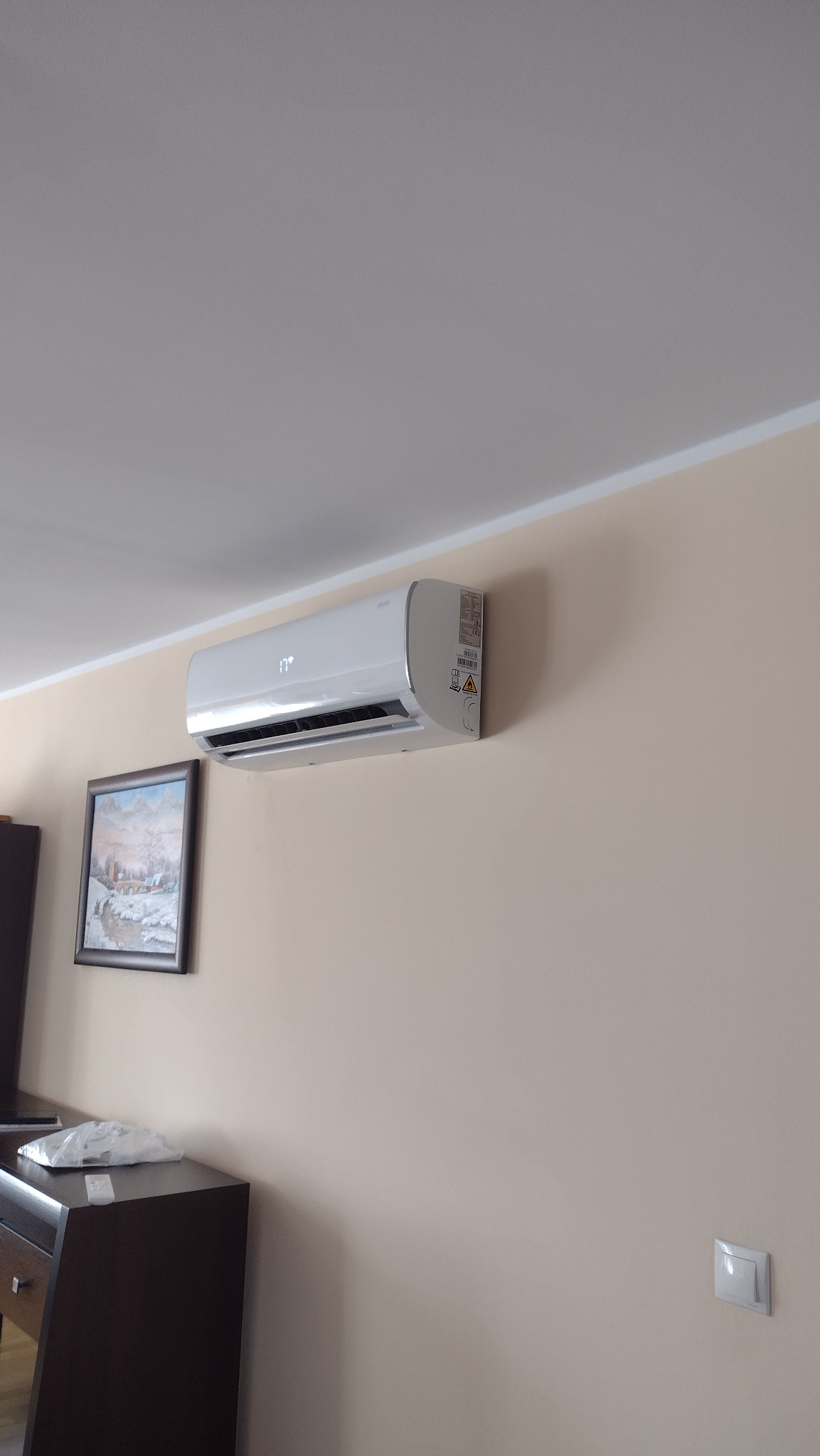 Biały klimatyzator ścienny zamontowany w pokoju, obok obrazu w ramie i włącznika światła, z widocznym wyświetlaczem temperatury.