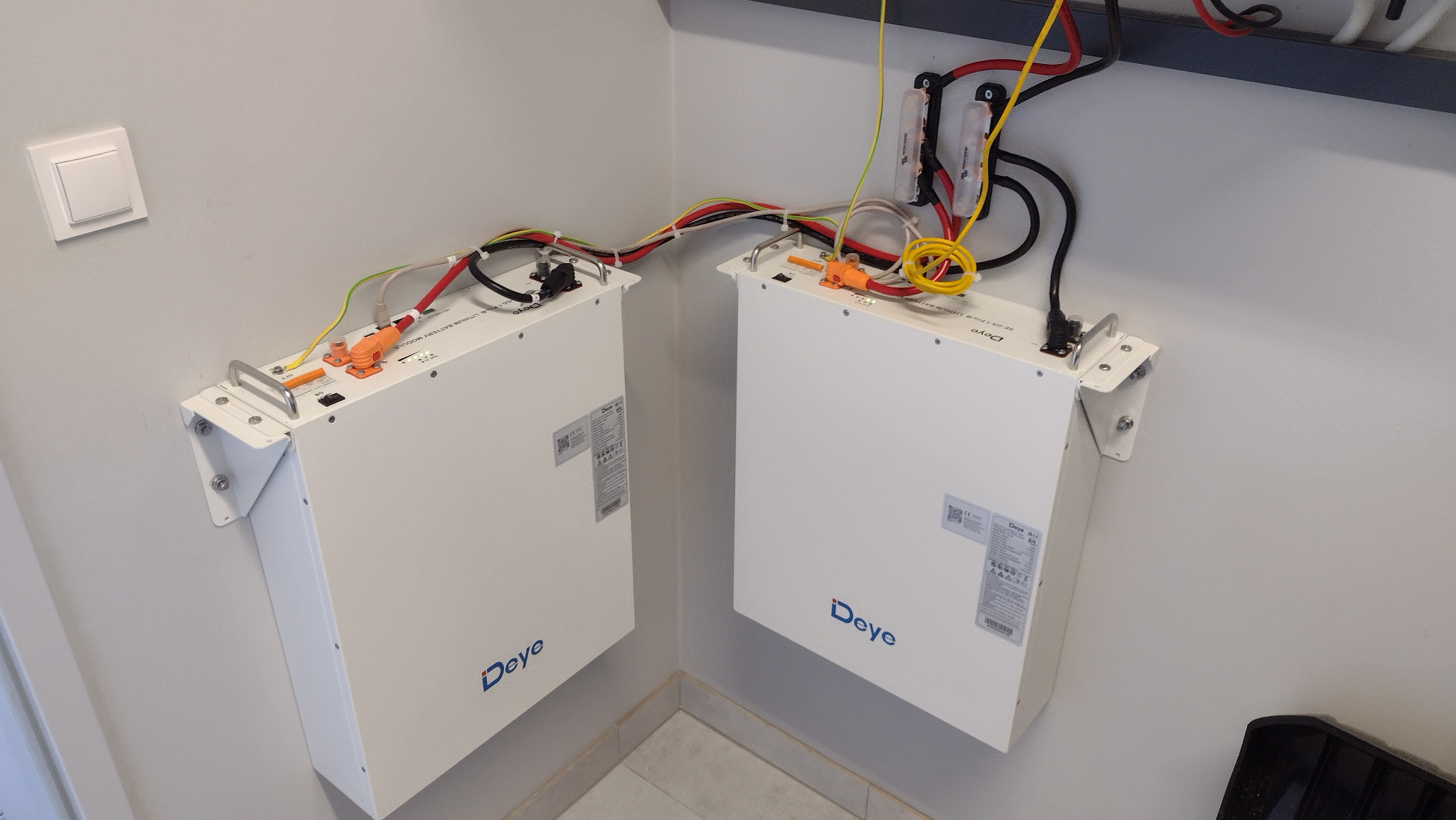 Dwa białe moduły magazynu energii Deye zamontowane na ścianie w rogu pomieszczenia, widoczne przewody i złącza.