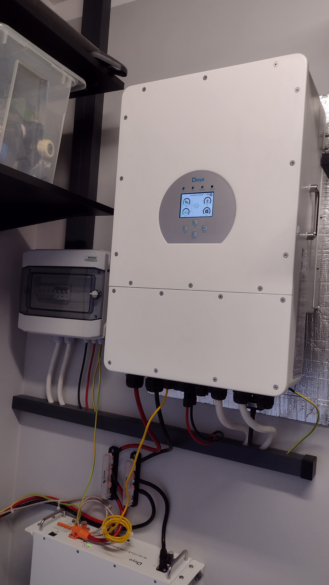 Biały inwerter solarny marki Deye zamontowany na ścianie, z widocznym panelem sterowania i podłączonymi kablami. Obok skrzynka bezpiecznikowa Noark i akumulator litowo-jonowy Deye. Widoczne...