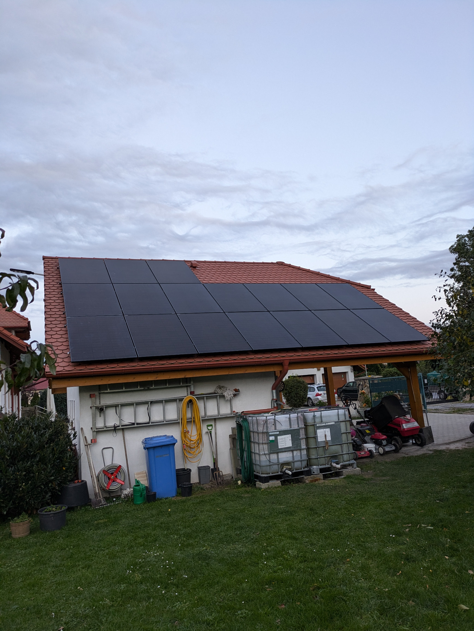 Panele fotowoltaiczne na dachu budynku gospodarczego z czerwoną dachówką, widok na ogród z narzędziami i zbiornikami na wodę.