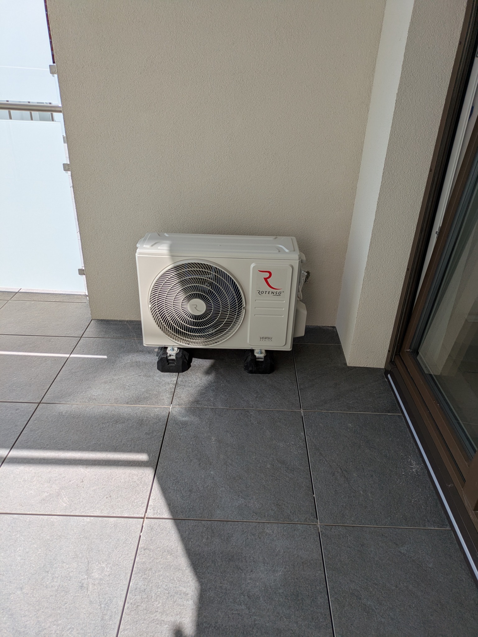 Biała jednostka zewnętrzna klimatyzacji marki Rotenso, model Versu Series, umieszczona na czarnych podkładkach antywibracyjnych na szarym balkonie.