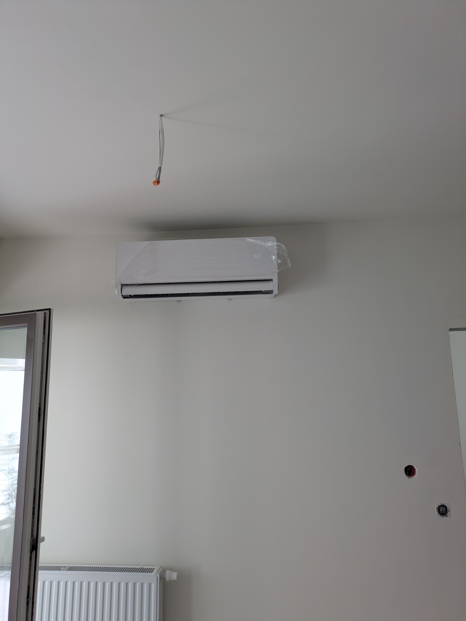 Nowoczesny, biały klimatyzator na ścianie, jeszcze z folią ochronną. Widoczne wystające kable ze ściany i grzejnik. Minimalistyczne wnętrze w jasnych barwach.