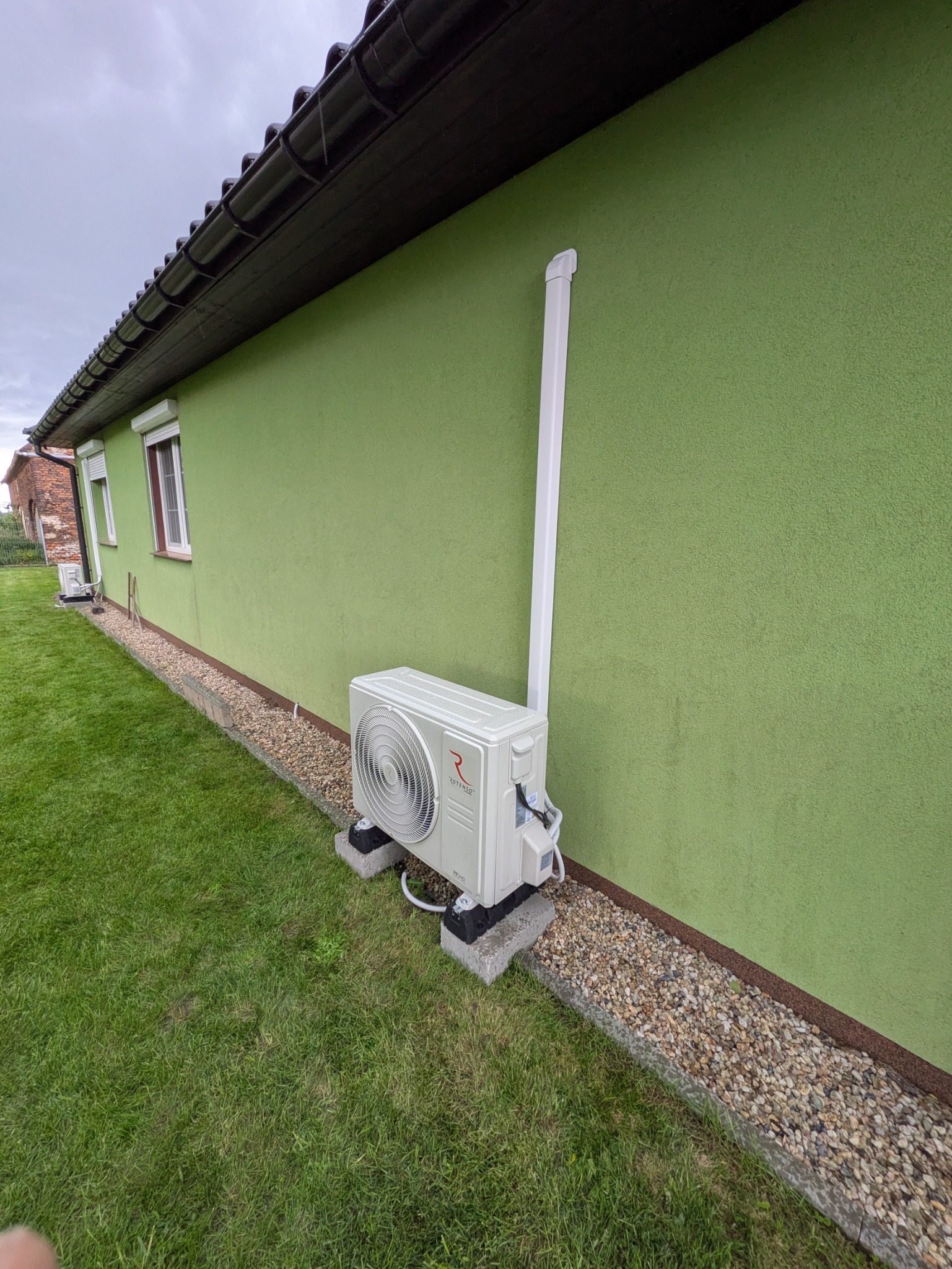 Zewnętrzna jednostka klimatyzacji Rotenso, zamontowana na betonowych podstawach przy zielonej ścianie domu. Widoczna instalacja odprowadzająca skropliny.