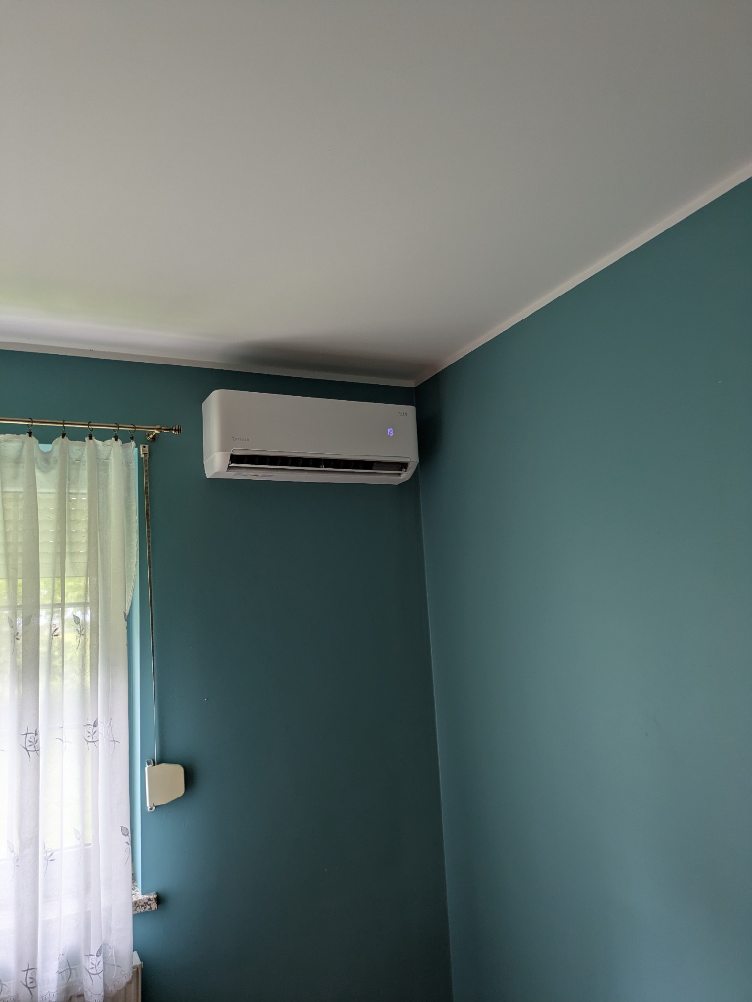 Klimatyzator Rotenso TETA zamontowany na ścianie w pokoju z turkusowymi ścianami, widoczne okno z zasłonami. Estetyczny montaż, nowoczesny design.