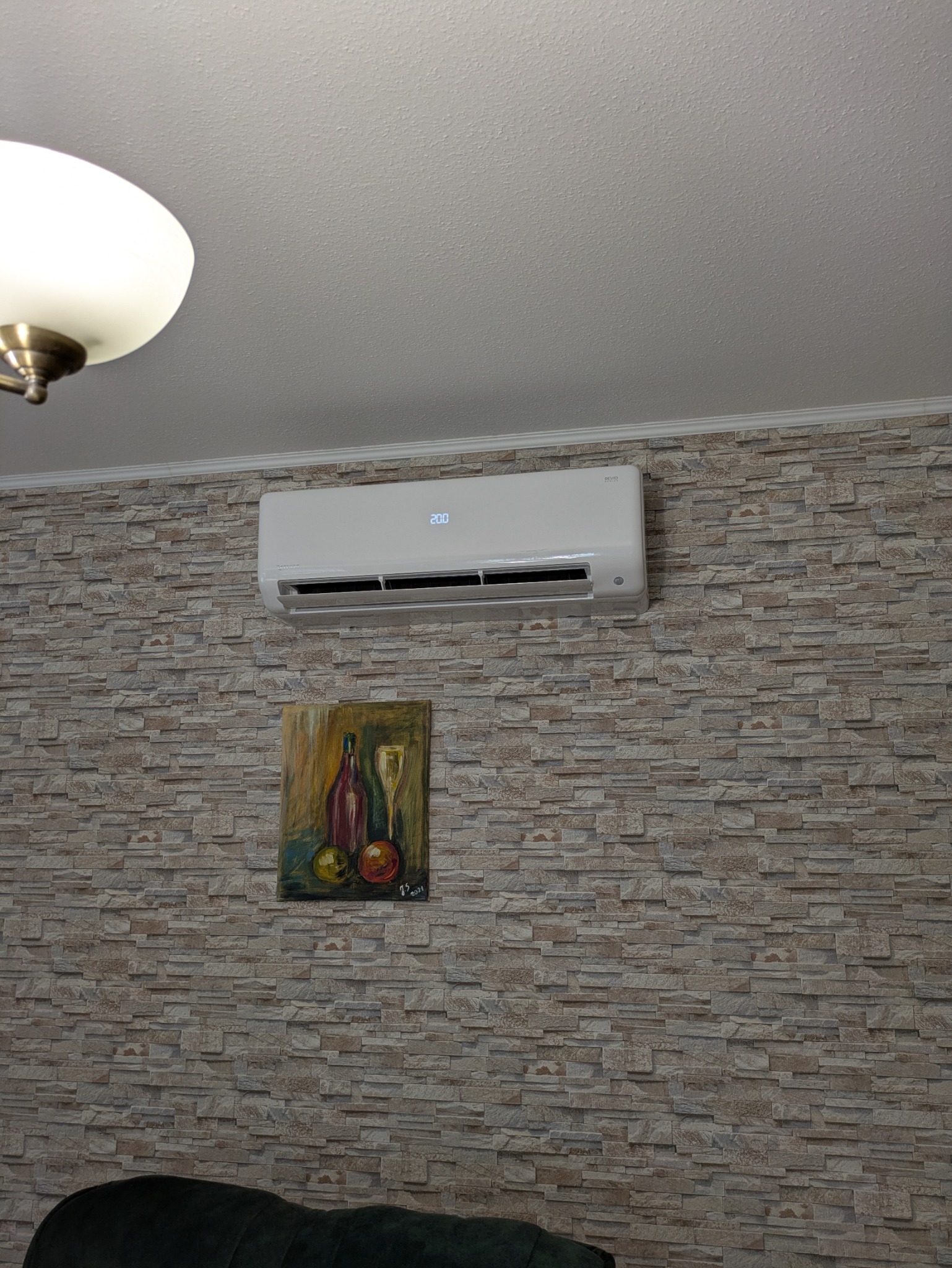 Biały klimatyzator na ścianie z dekoracyjnej cegły, pod nim obraz z motywem wina. Widoczny fragment lampy sufitowej i kanapy.