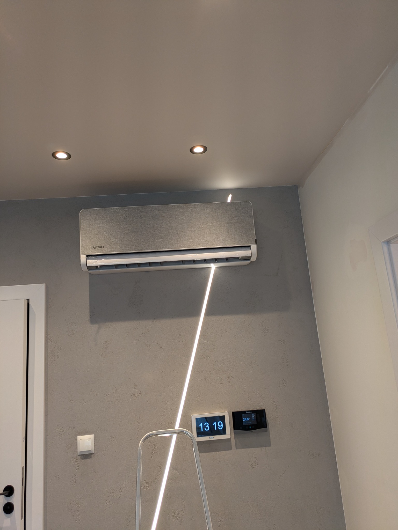 Szary klimatyzator Rotenso na ścianie o fakturze betonu, podświetlony ledową listwą. Widoczne panele sterowania i fragment drabiny.