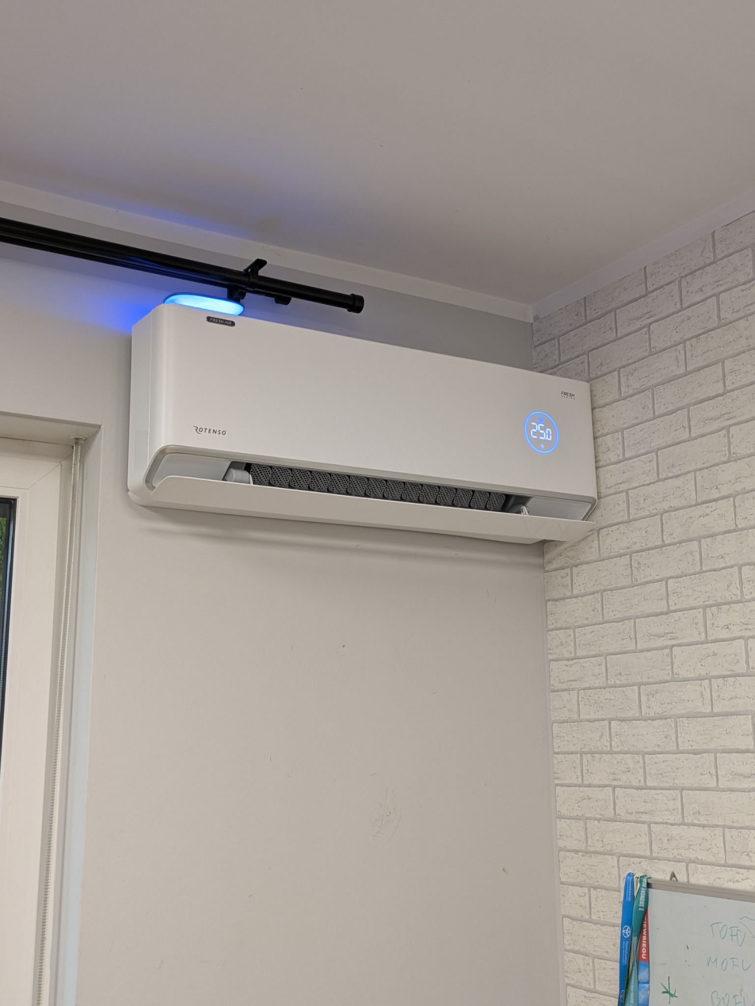 Biały klimatyzator Rotenso z niebieskim podświetleniem, zamontowany na szarej ścianie w pomieszczeniu z tapetą imitującą cegłę. Widoczny fragment okna i karnisz.