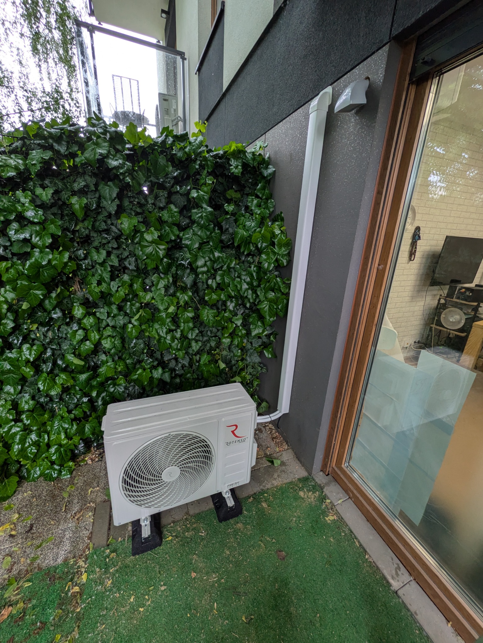 Biała jednostka zewnętrzna klimatyzacji Rotenso na zielonej trawie, obok ściany budynku z rurą odprowadzającą. Tło stanowi ściana z zielonego bluszczu.