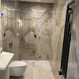 Wykończona łazienka z płytkami imitującymi marmur, wiszącą toaletą, grzejnikiem drabinkowym w kolorze czarnym oraz ukrytą szafką z lustrem.