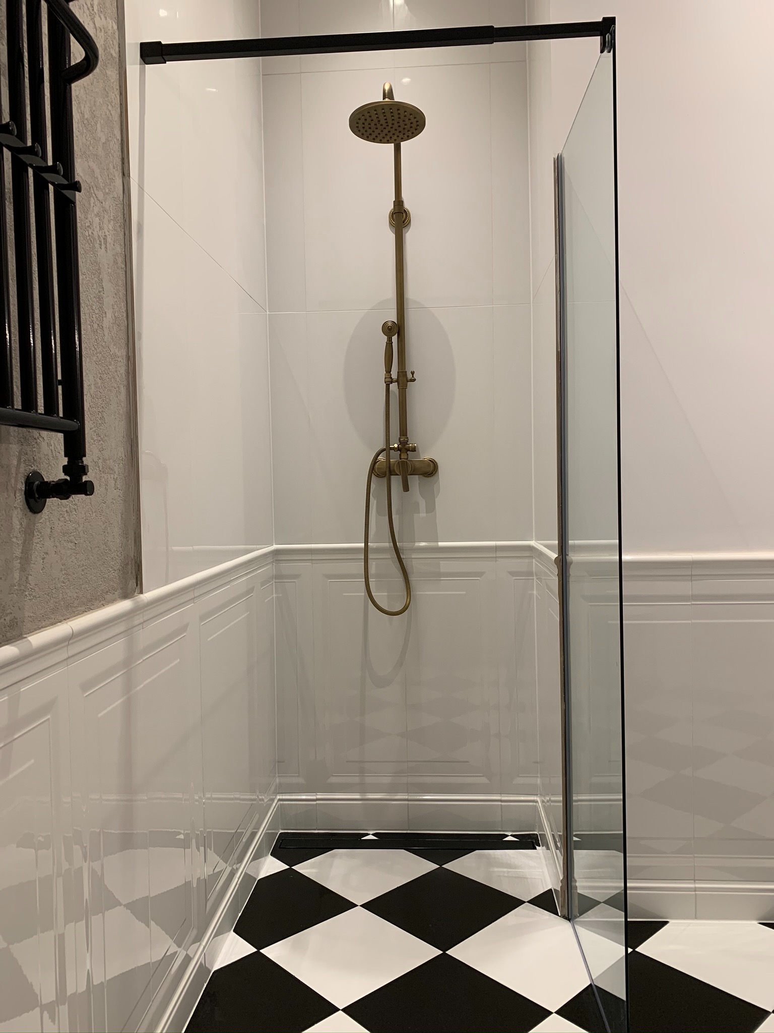 Kabina prysznicowa z deszczownicą w stylu retro, czarno-biała szachownica na podłodze, białe płytki ścienne z dekoracyjną listwą, grzejnik drabinkowy w kolorze czarnym.