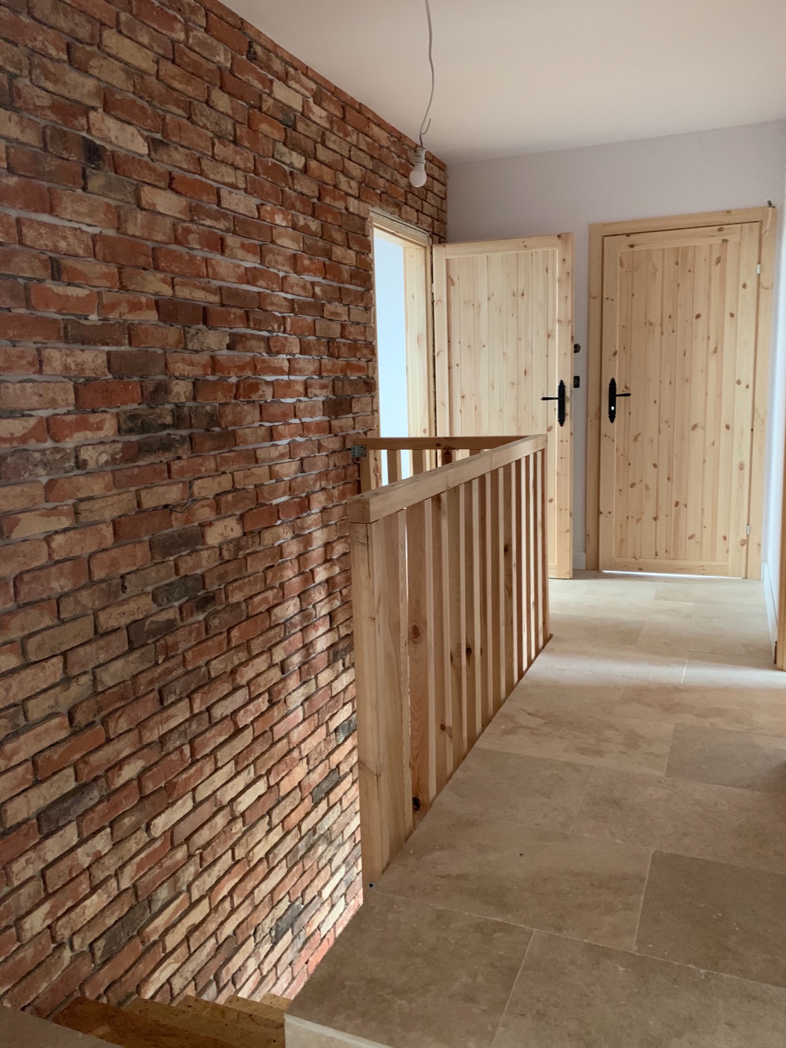 Wnętrze domu z ceglaną ścianą, drewnianą balustradą schodów i dwiema sosnowymi drzwiami z czarnymi klamkami.