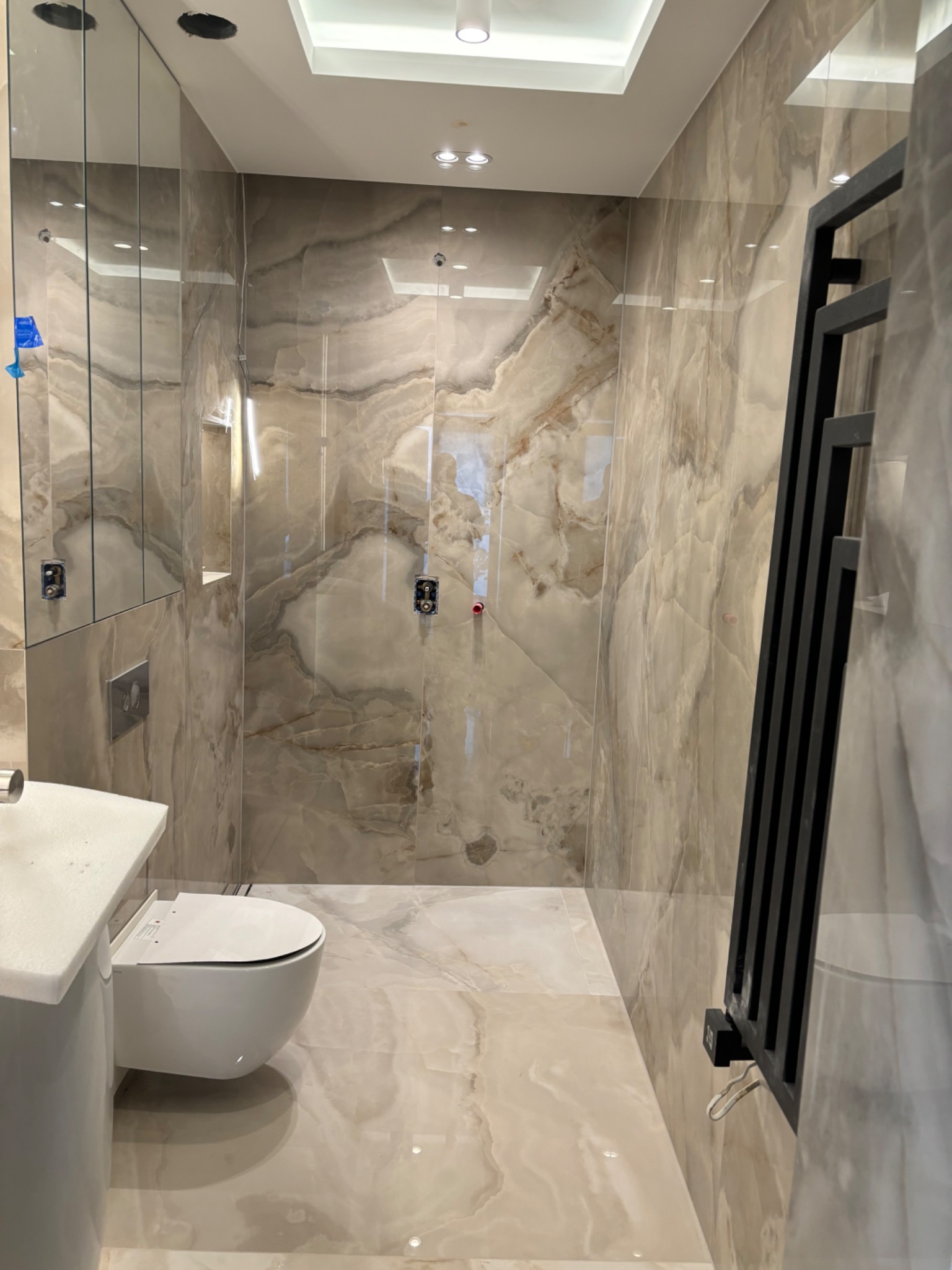 Wykończona łazienka z płytkami imitującymi marmur, wiszącą toaletą, grzejnikiem drabinkowym w kolorze czarnym oraz ukrytą szafką z lustrem.