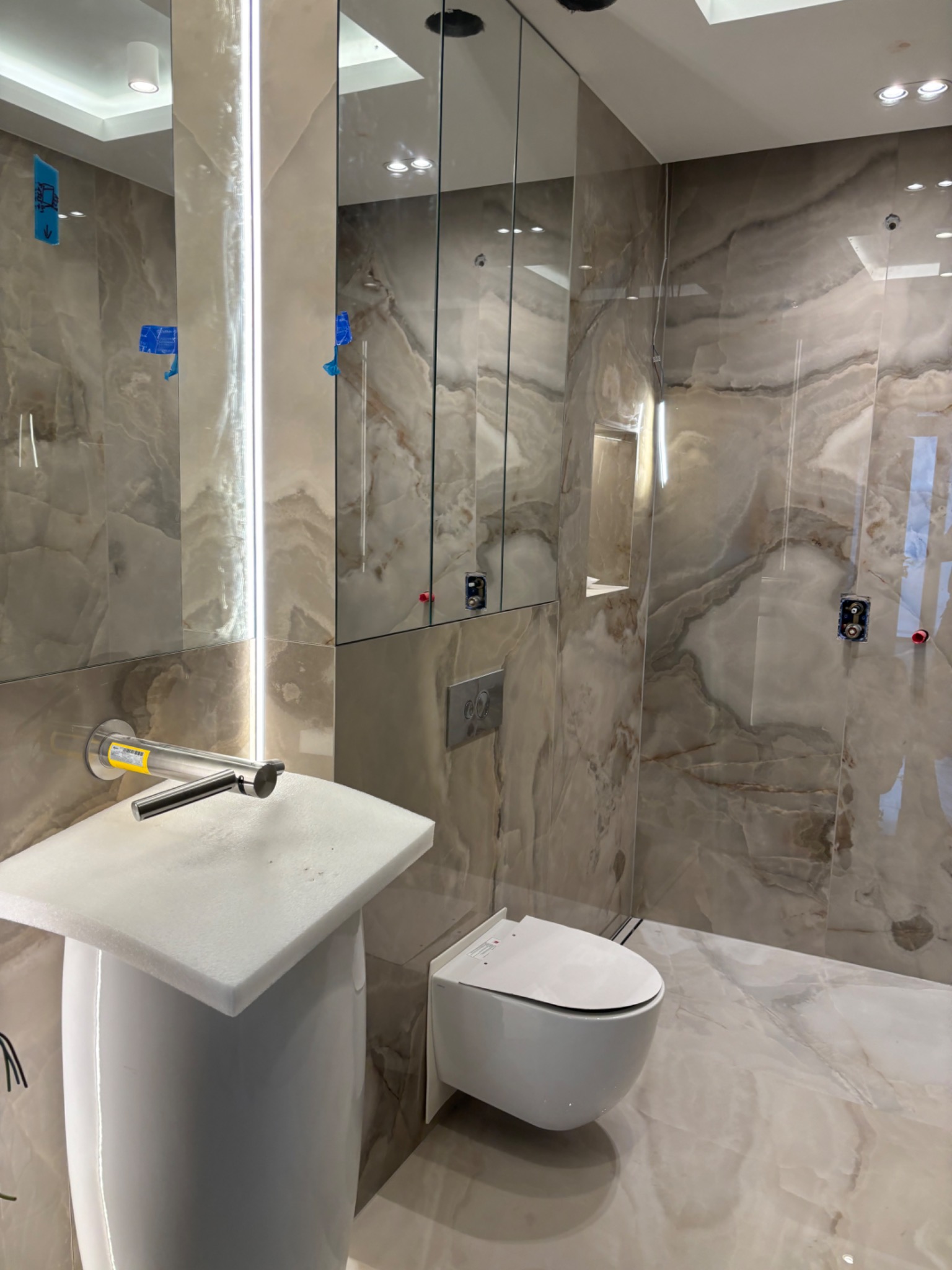 Nowoczesna łazienka z płytkami imitującymi marmur, wiszącą toaletą i umywalką na postumencie, widoczne niedokończone instalacje elektryczne.