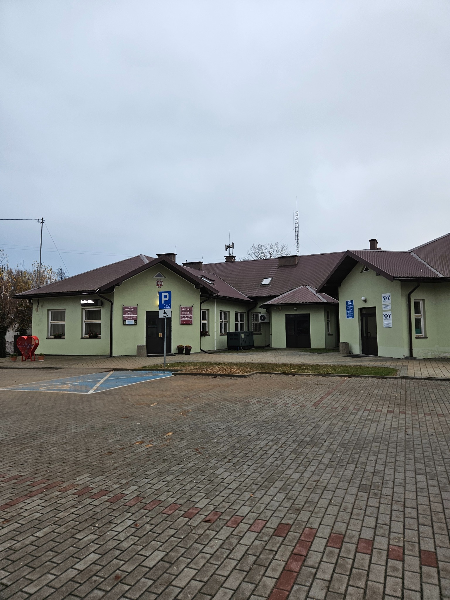 Budynek urzędu gminy z zieloną elewacją, brązowym dachem i utwardzonym placem przed wejściem. Widoczne tablice informacyjne i oznaczenie parkingu dla osób niepełnosprawnych.