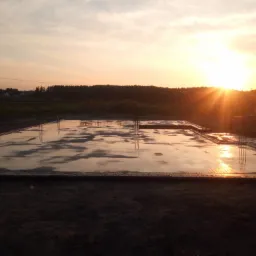 Zalana płyta fundamentowa z wystającymi prętami zbrojeniowymi odbijająca zachodzące słońce, w tle widoczna linia lasu i słup wysokiego napięcia.