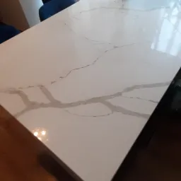 Biały stół z blatem o marmurkowym wzorze, na którym stoi wazon z białymi kwiatami, obok niebieskie krzesło.