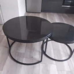 Dwa okrągłe stoliki kawowe z czarnymi blatami na metalowych nogach, większy i mniejszy, ustawione na panelach podłogowych w jasnym odcieniu drewna, w tle fragmenty szafek kuchennych.