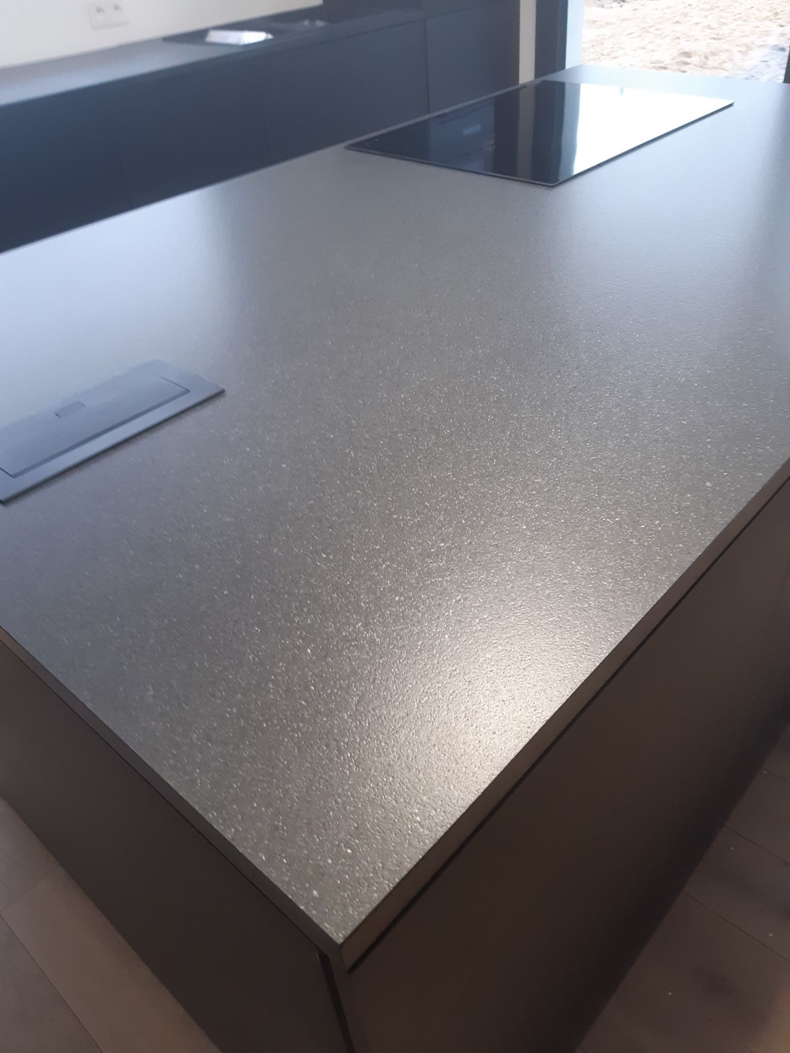 Szary, błyszczący blat kuchenny z wbudowaną płytą indukcyjną i gniazdem elektrycznym, zamontowany na ciemnych szafkach.
