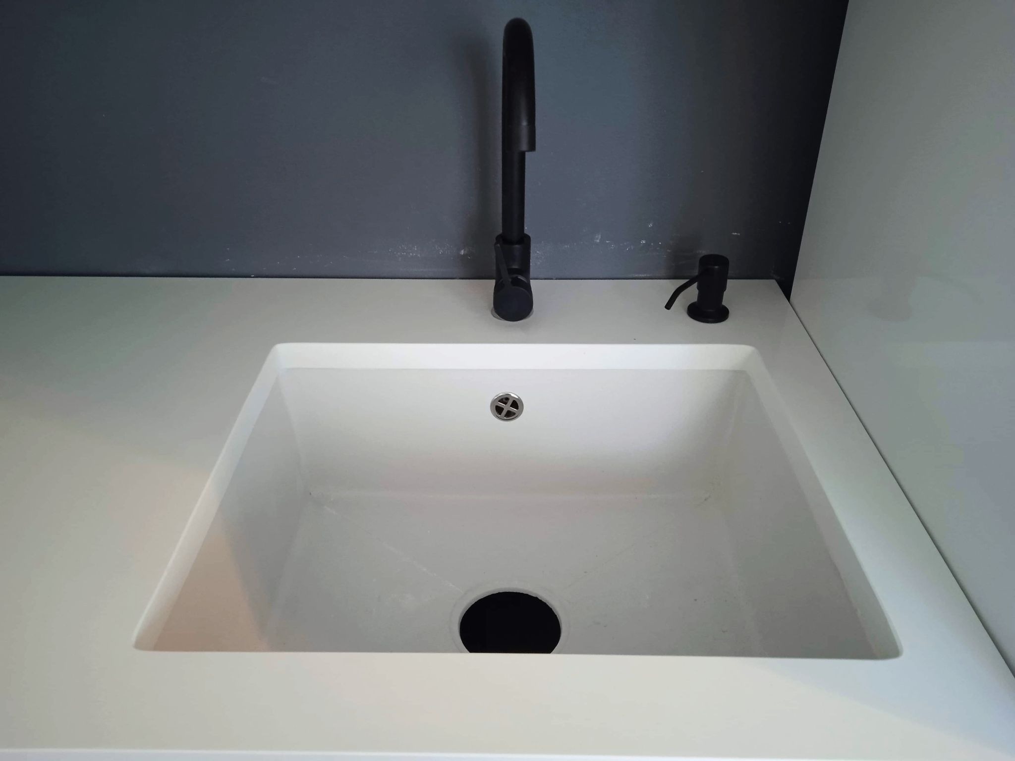 Biały zlew kuchenny z czarną baterią i dozownikiem mydła na tle szarej ściany.