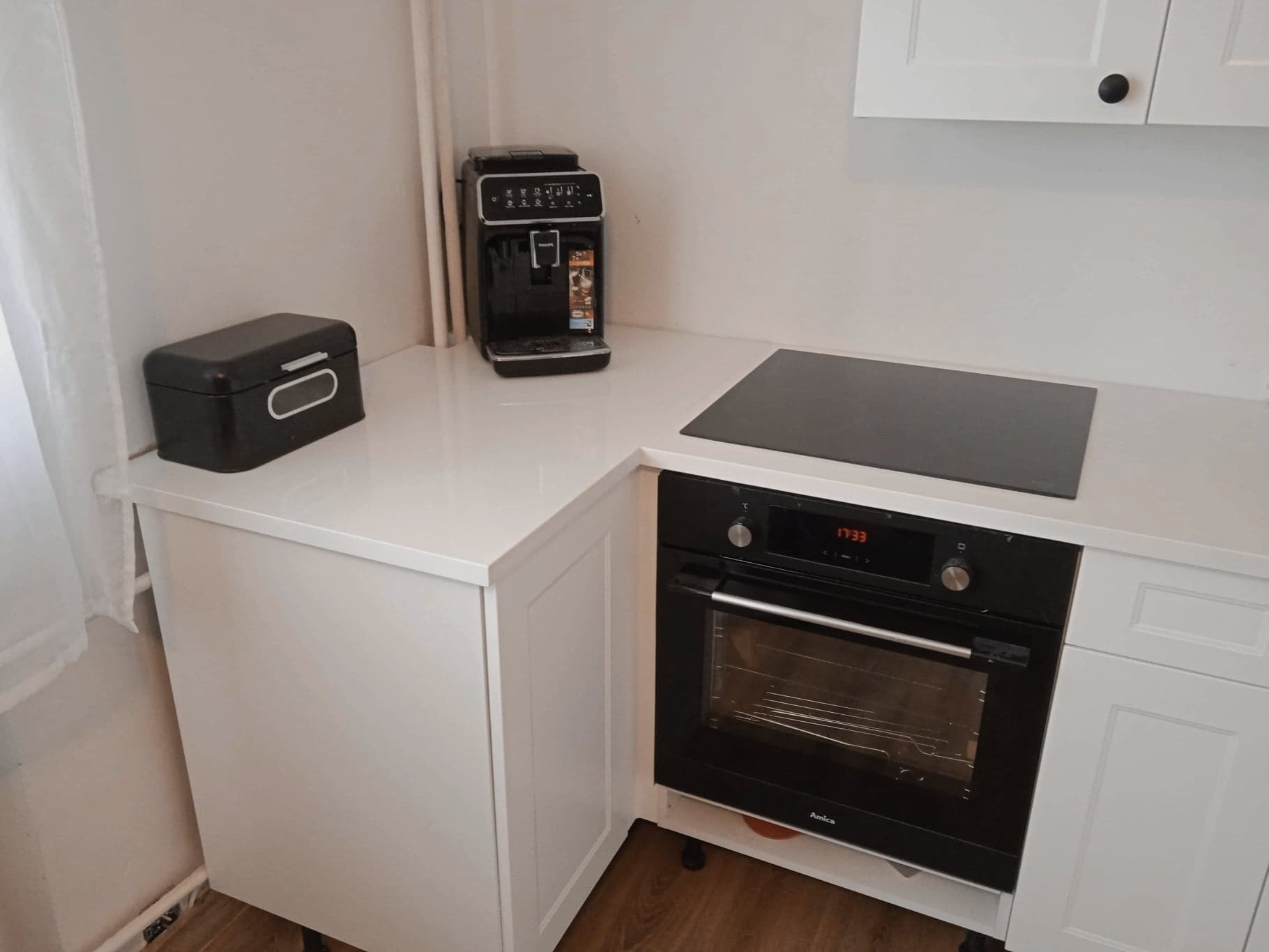 Biały, narożny blat kuchenny z automatem do kawy, chlebakiem i płytą indukcyjną nad piekarnikiem.