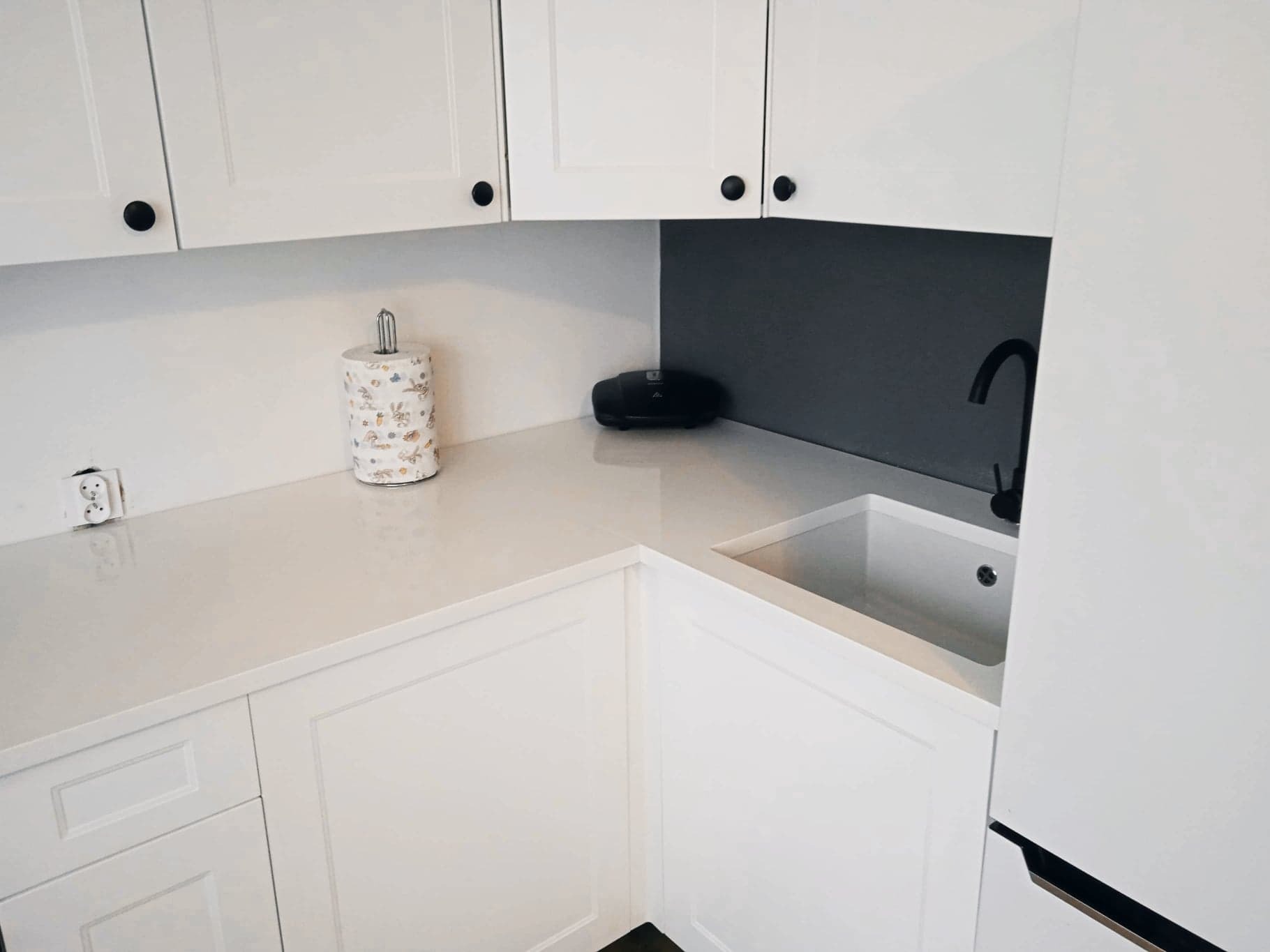 Jasny blat kamienny z czarną armaturą i białymi szafkami w kuchni w Szczecinie, widoczny dozownik na ręczniki papierowe.