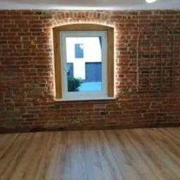 Ceglana ściana po renowacji z oknem i dekoracyjnym elementem imitującym okno, drewniana podłoga, widoczne ślady po usunięciu starej fugi i uzupełnieniu nową, ściana pomalowana lakierem do cegły.