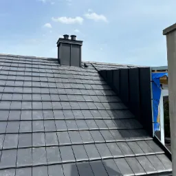 Nowy dach z ciemnej dachówki ceramicznej z kominem w kolorze antracytowym, widok z góry, częściowo widoczna ściana budynku w trakcie budowy.