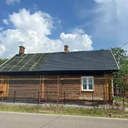 Drewniany dom z dachem w trakcie częściowej wymiany: połowa dachu pokryta czarną dachówką, druga połowa z widoczną konstrukcją więźby dachowej, rusztowania przy ścianie