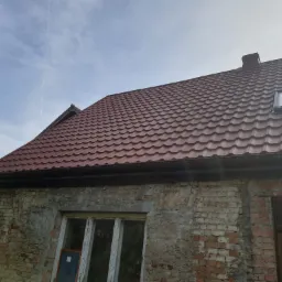 Czerwony dach z dachówką karpiówką na starym, ceglanym budynku z widocznymi śladami napraw, czarne rynny, okno dachowe i komin.