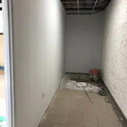 Wnętrze w trakcie prac wykończeniowych: świeżo pomalowana ściana w kolorze złamanej bieli kontrastuje z surowym sufitem podwieszanym i ścianą o strukturze baranka, na podłodze widoczne narzędzia...